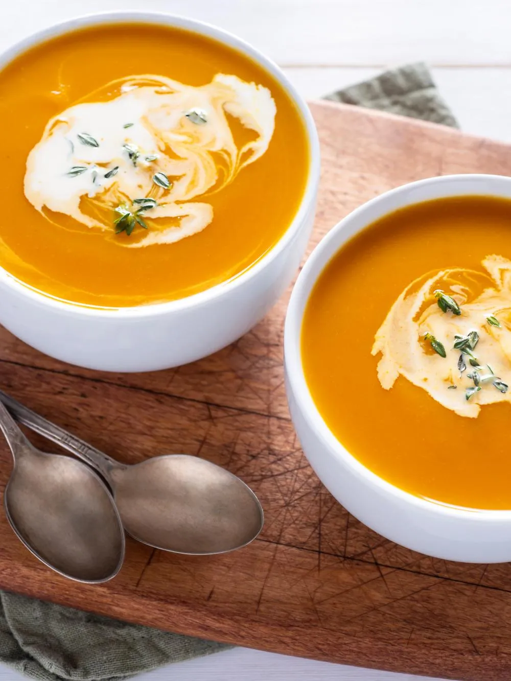 two white bowls filled with butternut squash soup and swired with coconut cream. They are sitting atop a cutting board with two silver spoons