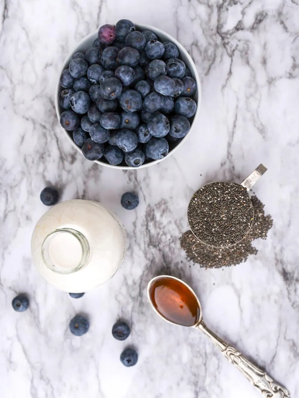 a marble table top with a bowl of blueberries, a measuring cup of chia seeds, a small jar of almond milk and a tablespoon of honey