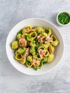 a white bowl of dairy free salmon pasta with peas and a small white bowl of peas sitting to the side of it/