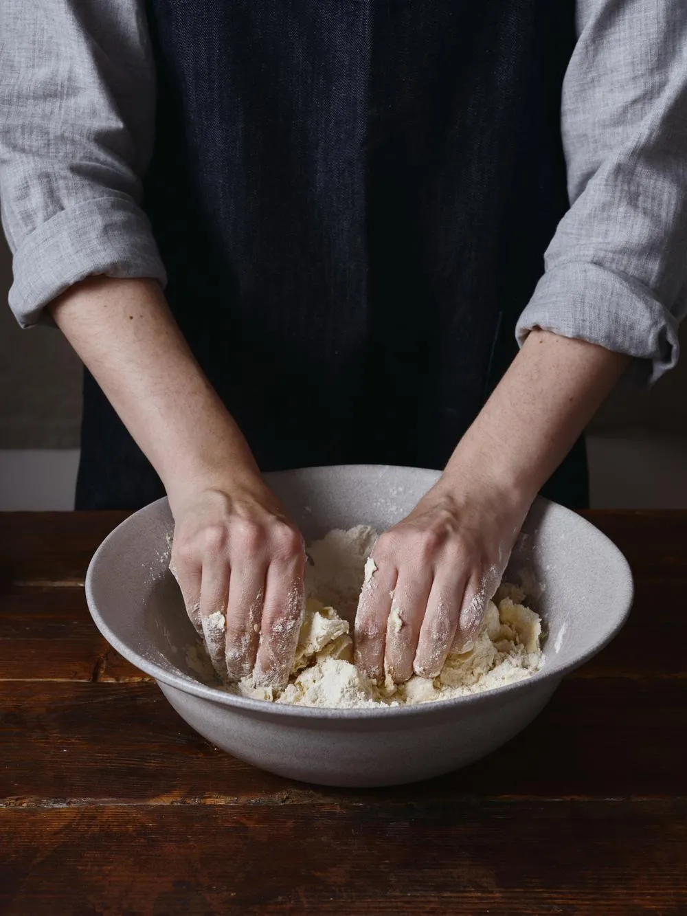a woman in a black apron with her hands in a white bowl kneading dough