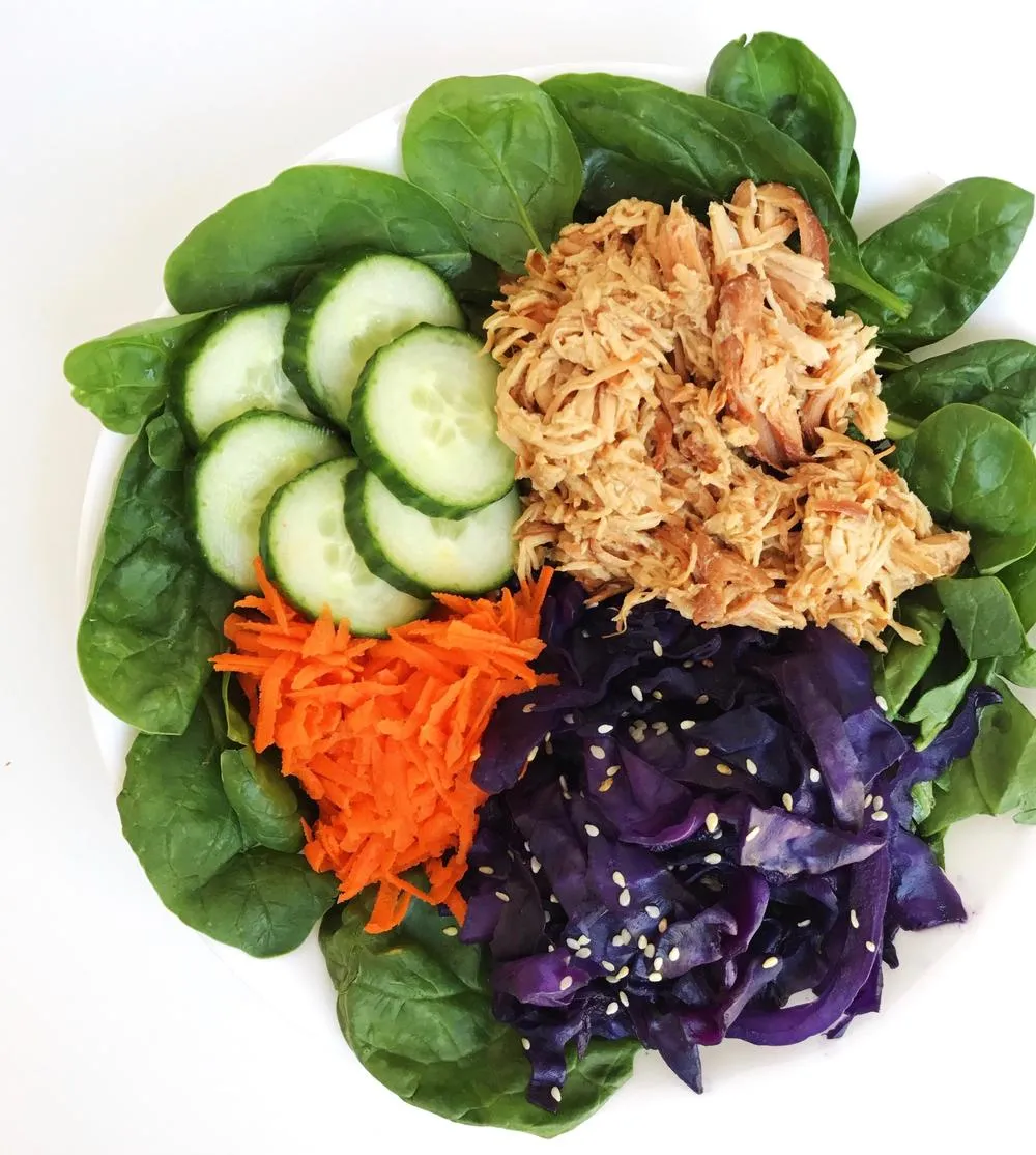 a white bowl full of spinach, shredded carrots, purple cabbage, sliced cucumbers, and slow cooker Asian honey chicken