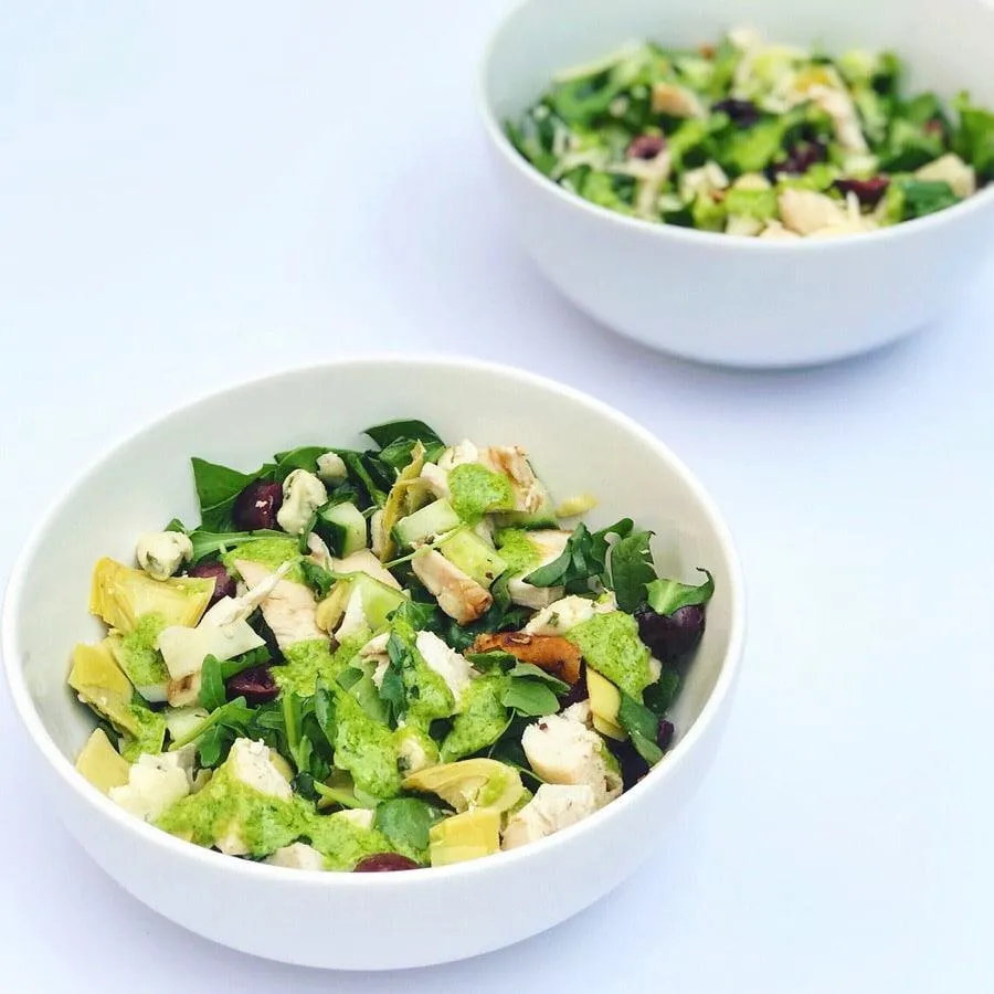 two salad bowls full of arugula, artichoke hearts, chicken, olives, cucumber, and basil dressing