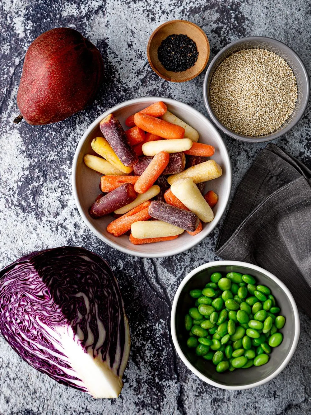 A marble table with bowls of carrots and beans surrounded by other fruits, vegetables, and grains