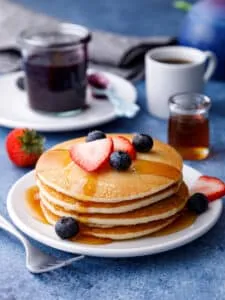 Stack of almond milk pancakes on a table with syrup, blueberry compote and a mug of hot tea on a table with a strawberry