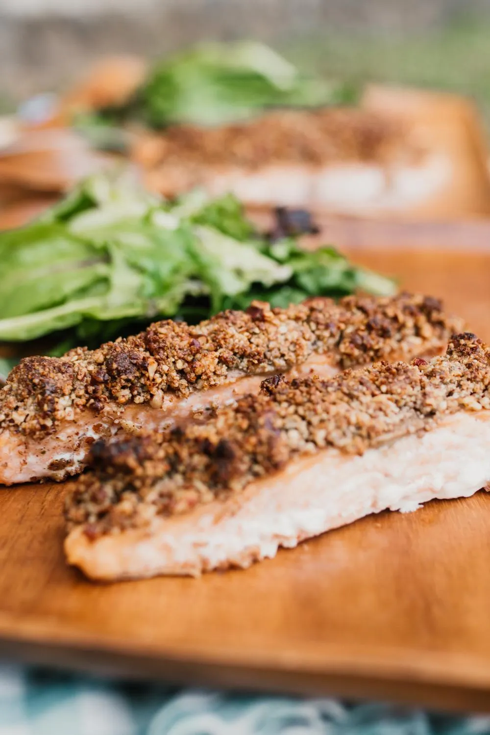 a checkered blue and white napkin with a wooden plate that has two pieces of honey pecan crusted salmon with a small side salad
