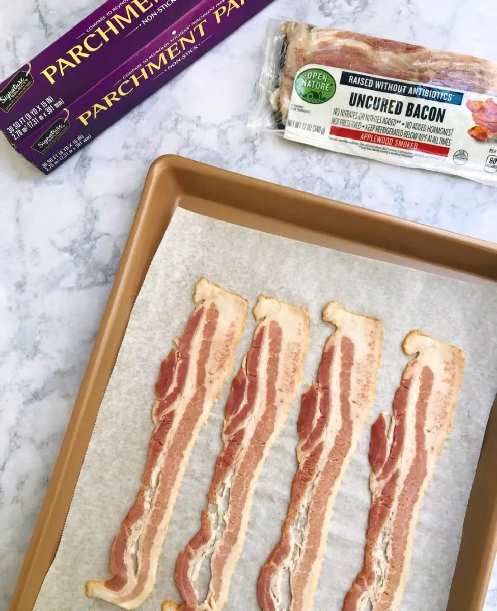 Four strips of bacon layed out on a baking sheet lined with parchment paper. Parchment paper and bacon in the background.