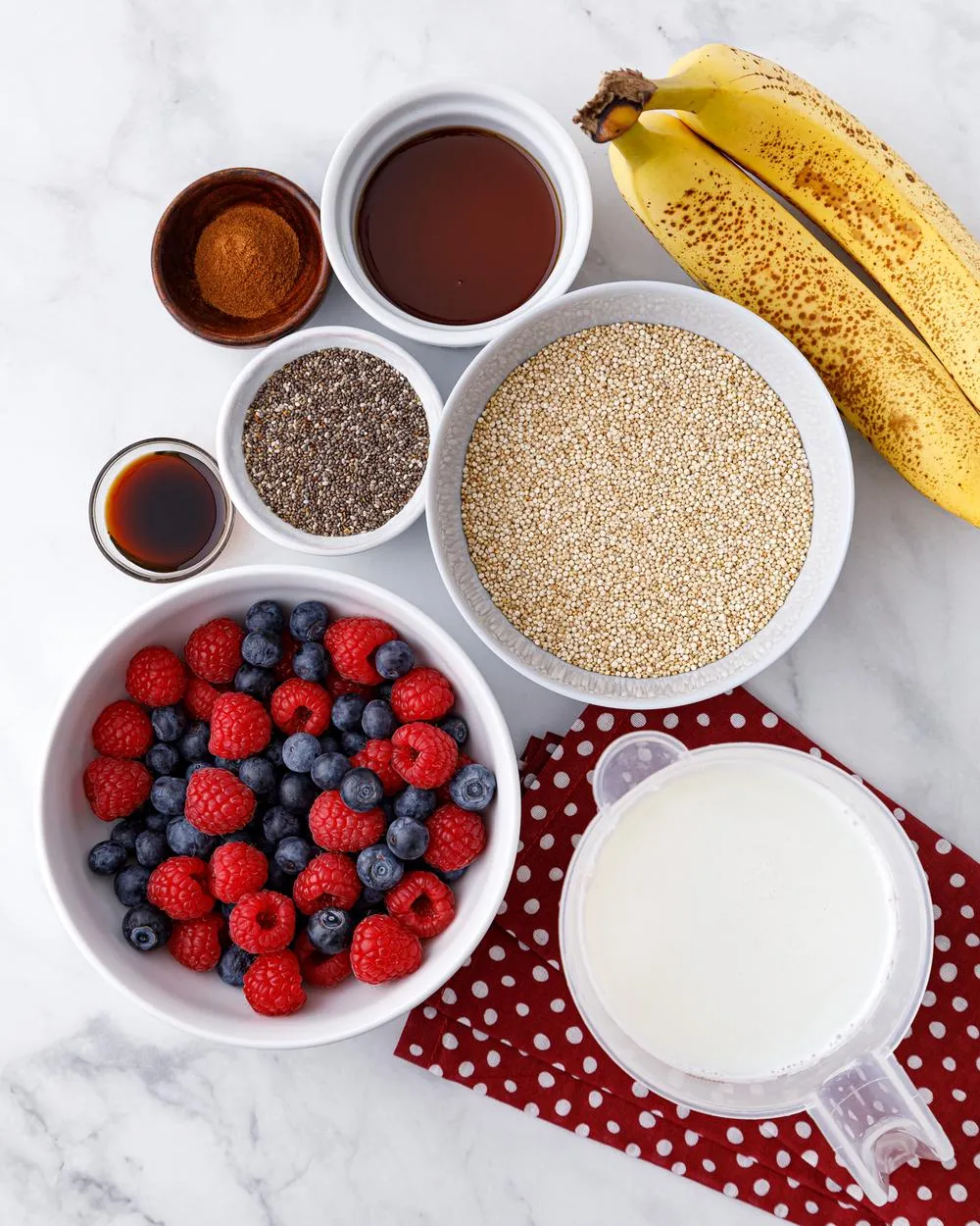 a white bowl of fresh blueberries and raspberries next to a measuring cup of plant milk, a white bowl of dry quinoa, and small containers filled with maple syrup, cinnamon and chia seeds, plus two ripe bananas all sitting on a white marble tabletop
