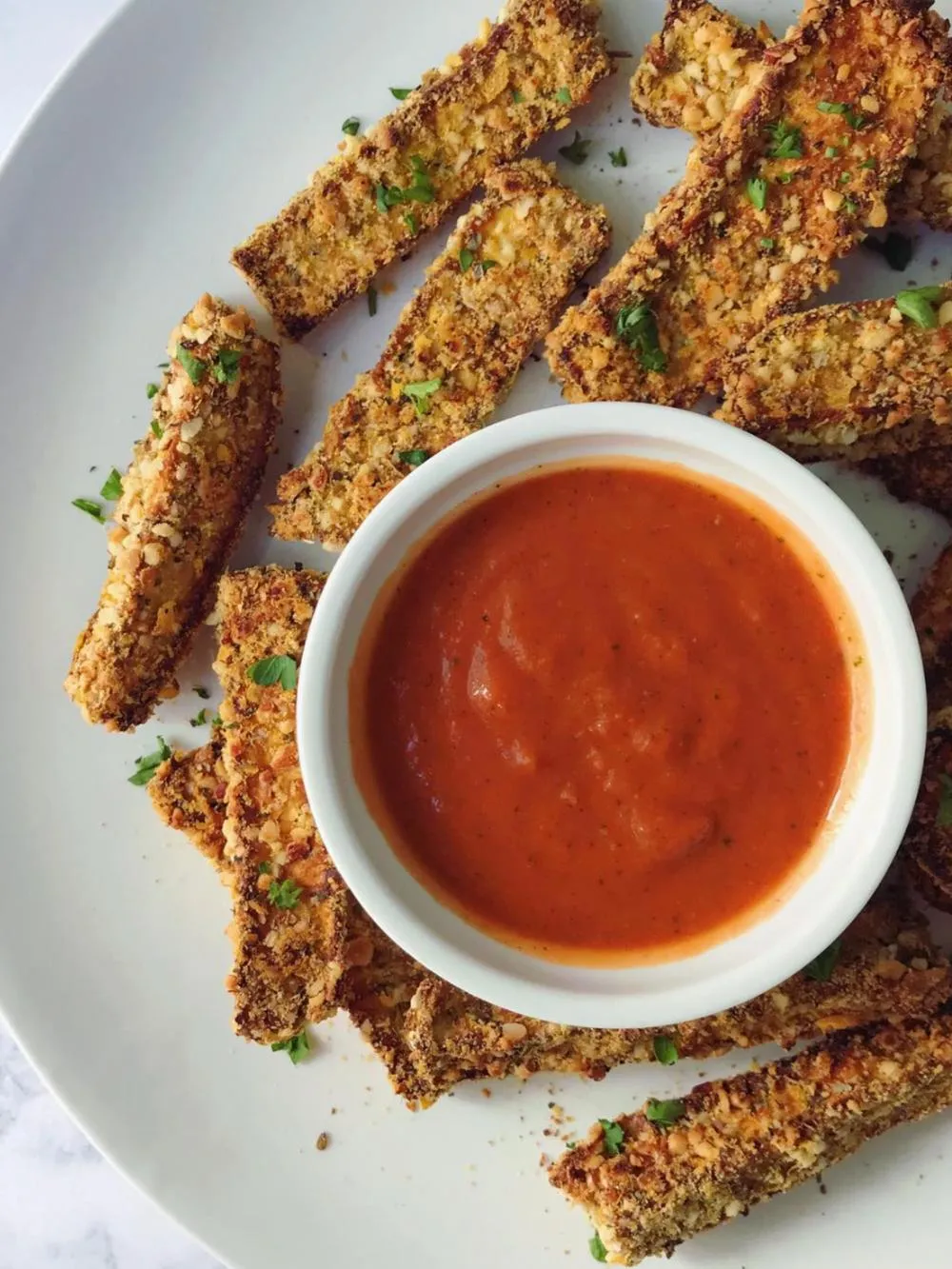 a white plate with crispy eggplant fries and a small white bowl of no tomato sauce on a white table