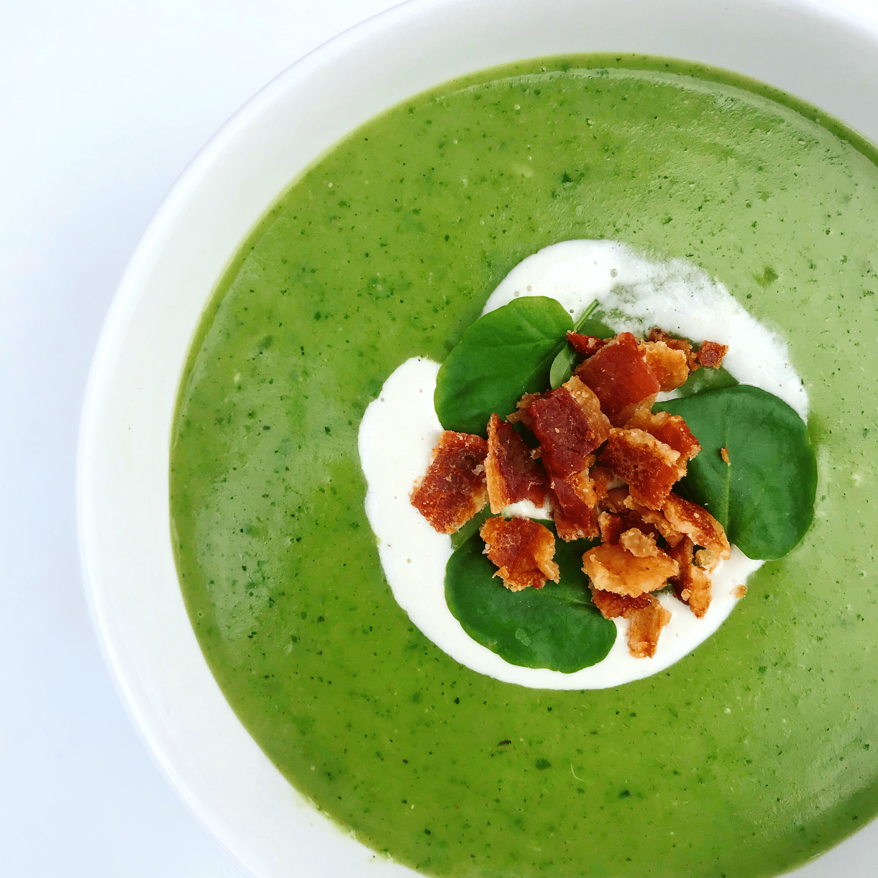 white bowl full of vibrant green zucchini watercress soup, topped with cream, bacon, and watercress leaves
