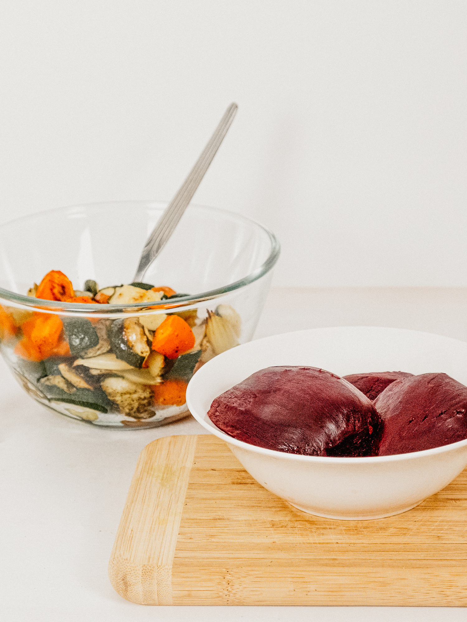 Roasted Beets in a white bowl on a cutting board next to a clear glass bowl of roasted carrots, zucchini, celery and turnips with a silver spoon