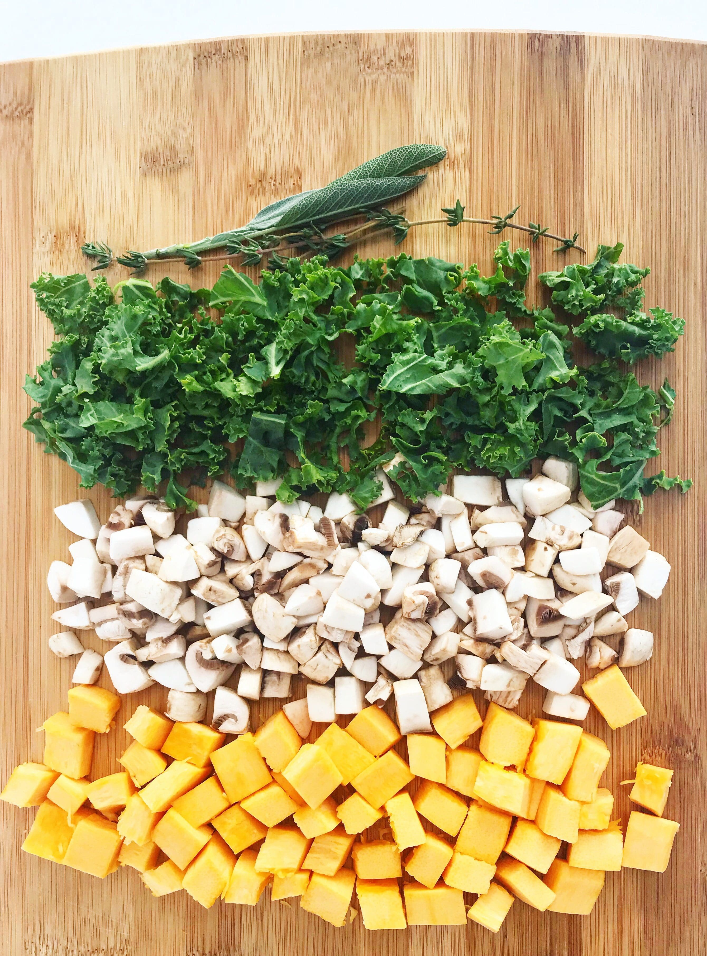Wooden cutting board with cut up pumpkin, mushrooms, kale, and fresh herbs.