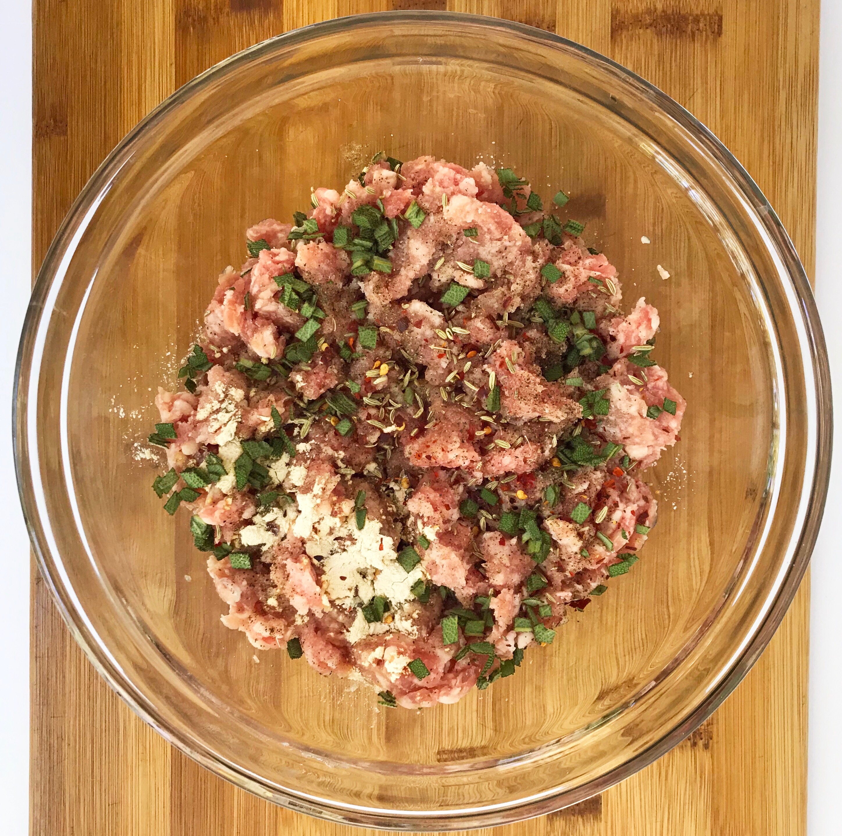 Preparation shot of a mixing bowl with ground pork, seasonings, maple syrup, and fresh sage on a wooden cutting board.
