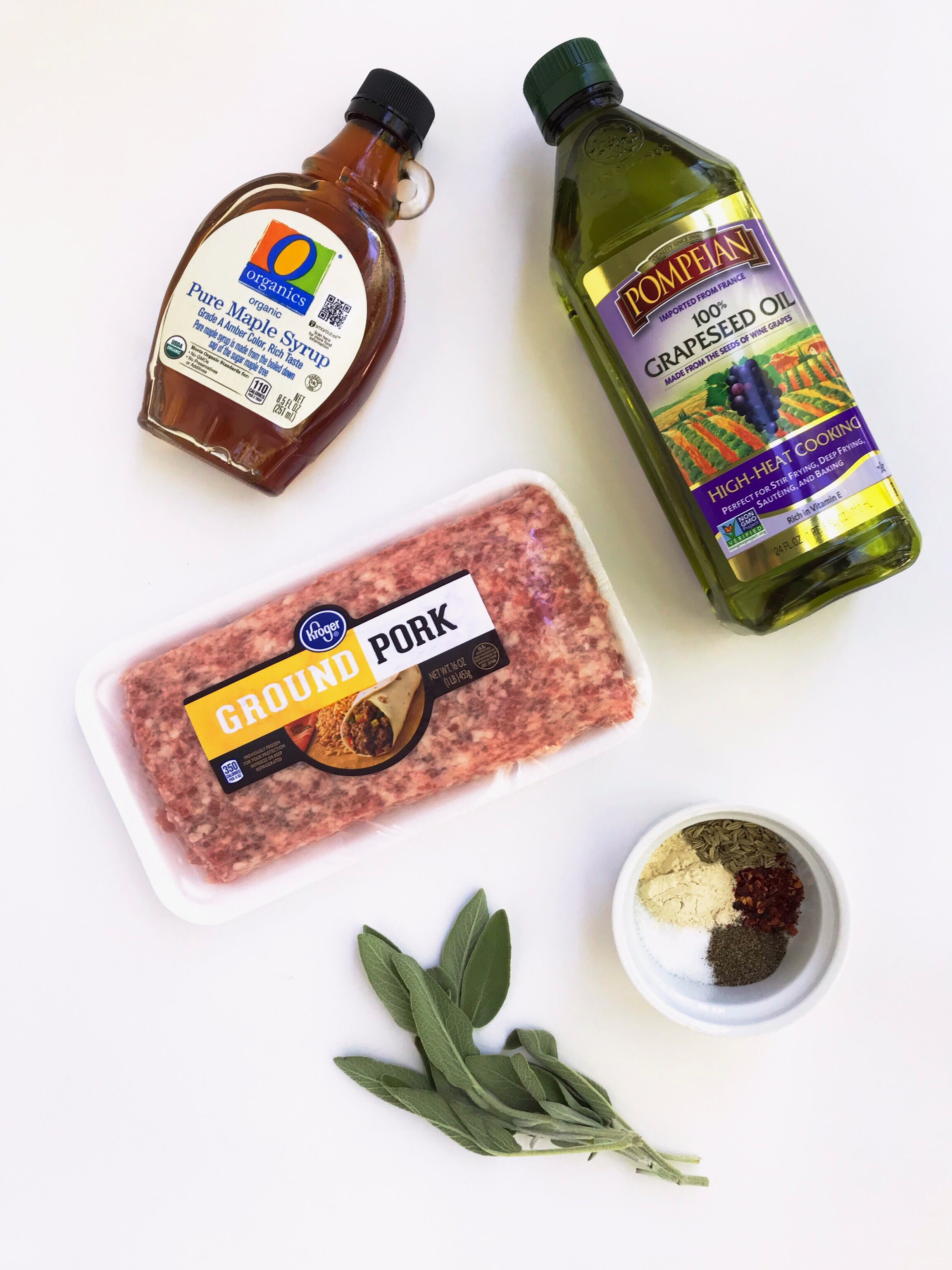A spread of the ingredients needed to make Maple-Sage Breakfast Patties. Ingredients displayed are ground pork, maple syrup, fresh sage, grapeseed oil, and a mix of dry seasoning.