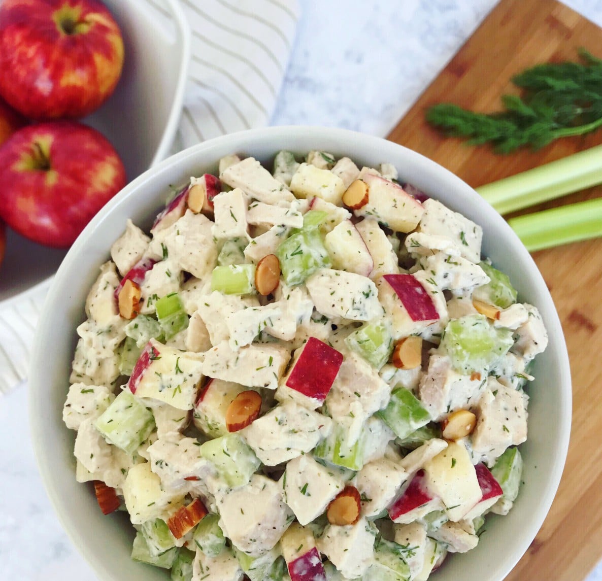 A bowl of Fresh Dill Chicken Salad, with apples, fresh dill, celery, and a cutting board in the background