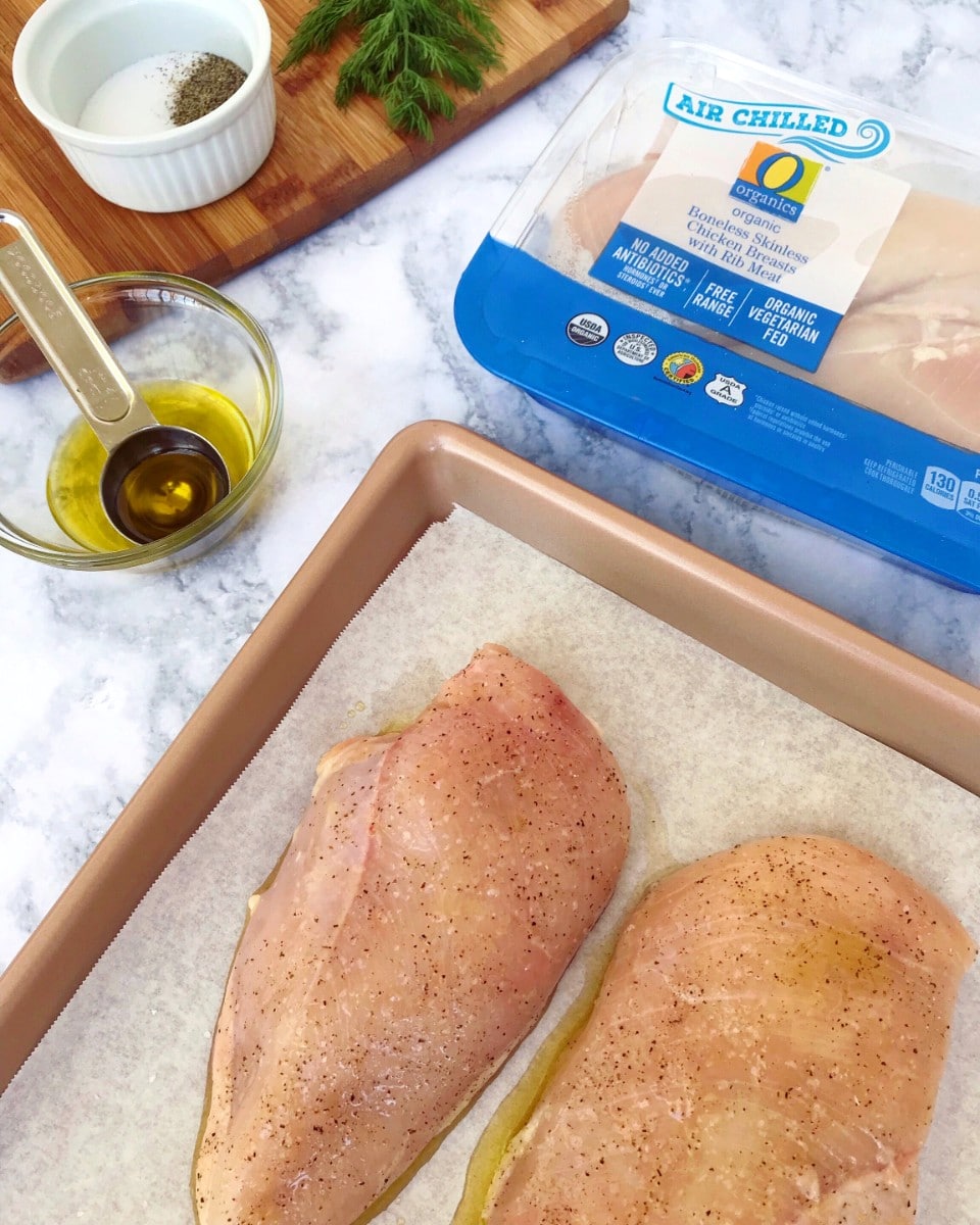 Air Chilled O Organics Chicken Breasts on a baking sheet, with a small bowl of olive oil, spices, fresh dill, and cutting board in the background