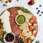 A gluten-free and dairy-free charcuterie board displaying meat, cheese, nuts, fruit, fresh herbs, crackers, olives, and pickles.