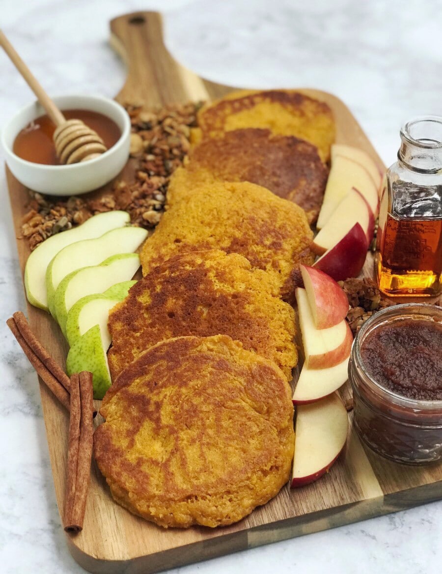 A wooden board with pancakes, apple and pear slices, maple syrup, honey, apple butter, granola, and pumpkin seeds