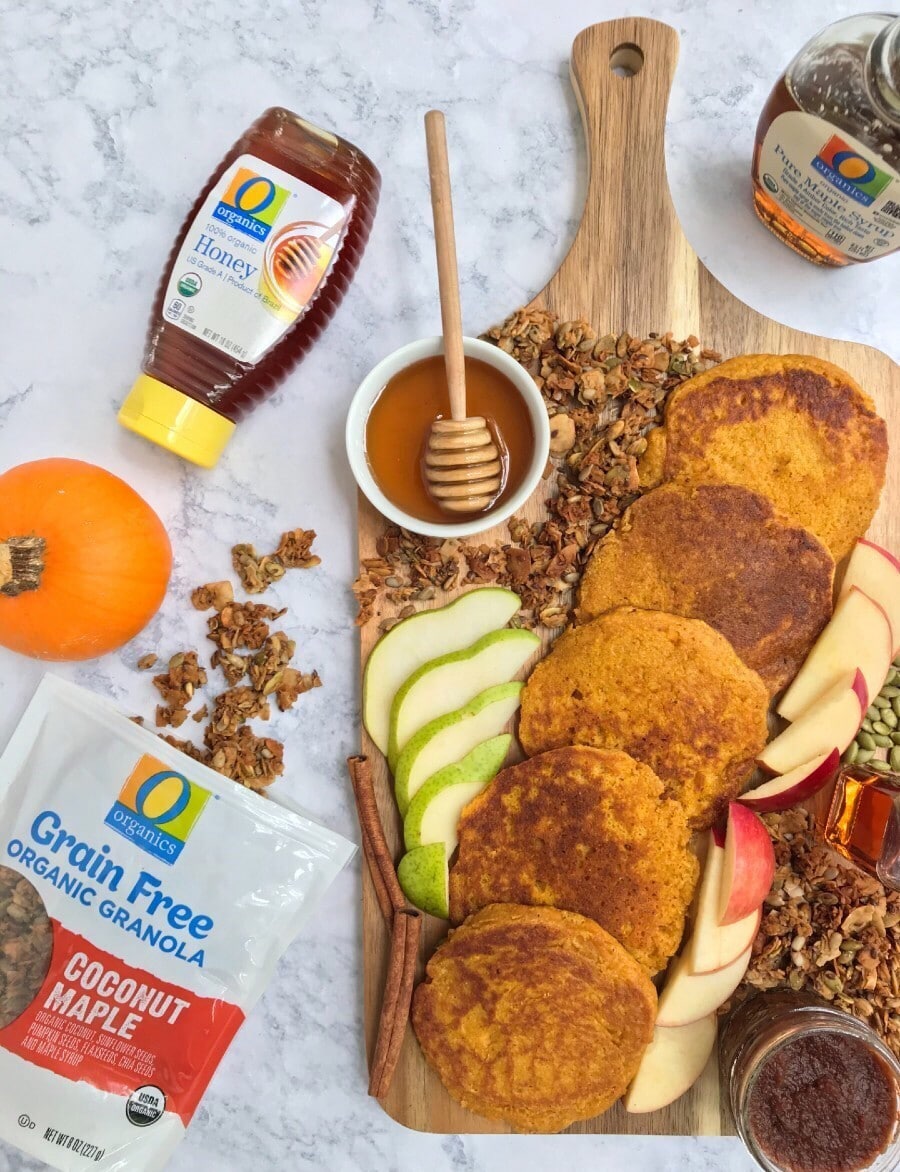 A wooden board with pancakes, apple and pear slices, maple syrup, honey, apple butter, granola, and pumpkin seeds. Also, featured is O Organics honey, maple syrup, and grain-free granola.