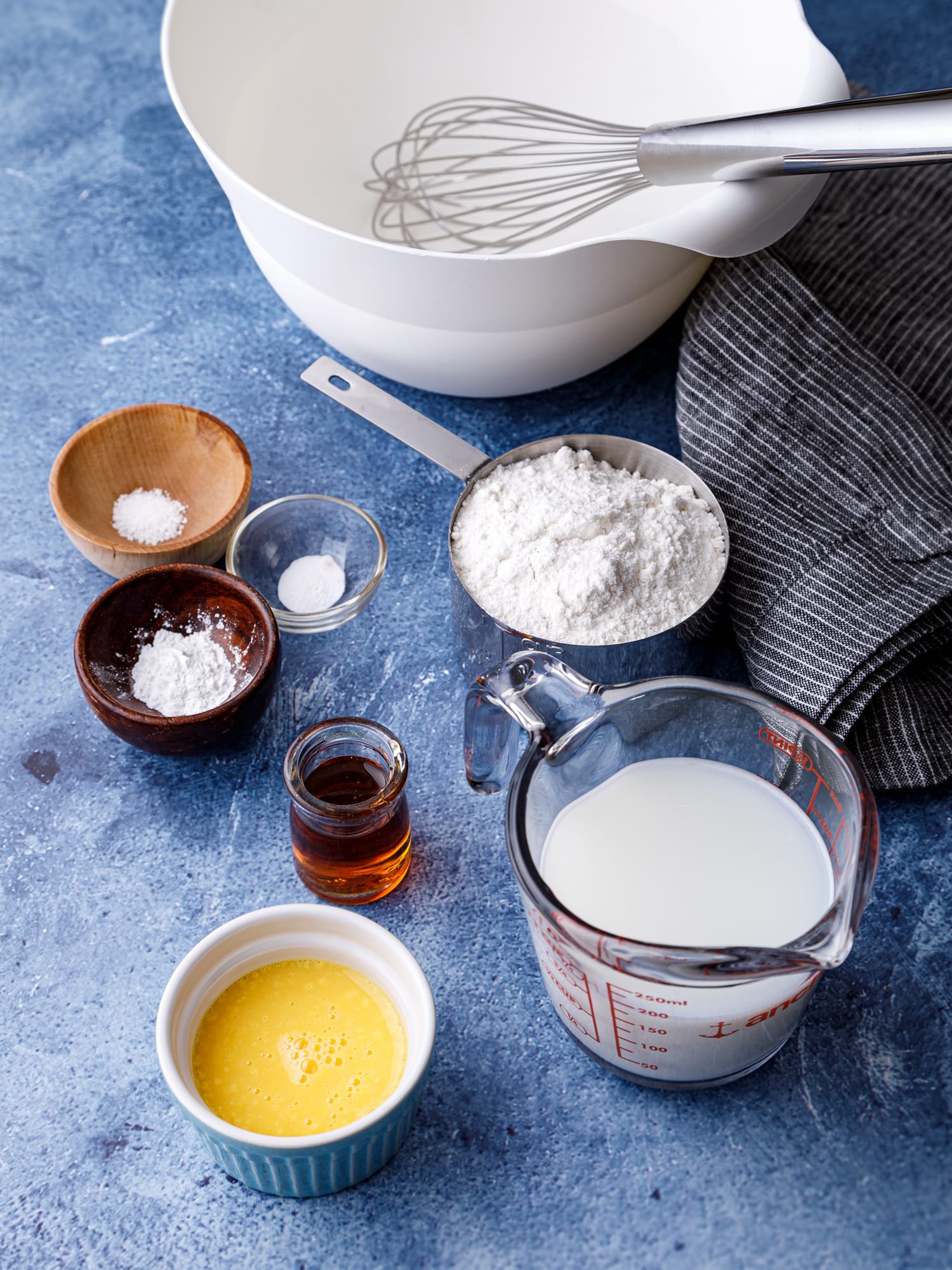 White bowl with a whisk a measuring cup of gluten free flour and almond milk a bowl of egg substitute, vanilla and baking soda on a table with a dishcloth