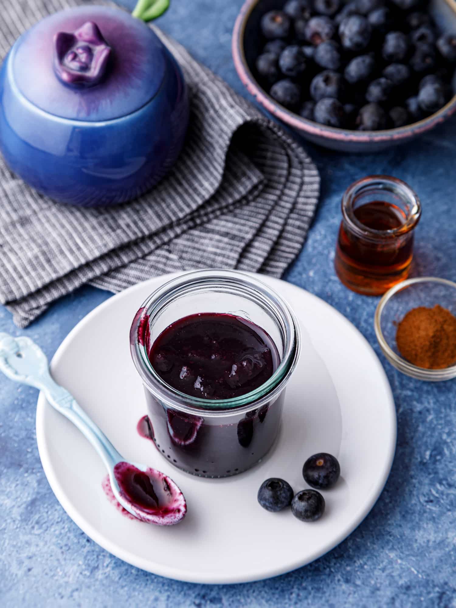 jar of homemade blueberry cinnamon sauce on a plate next to a bowl of fresh blueberries a blue serving pot