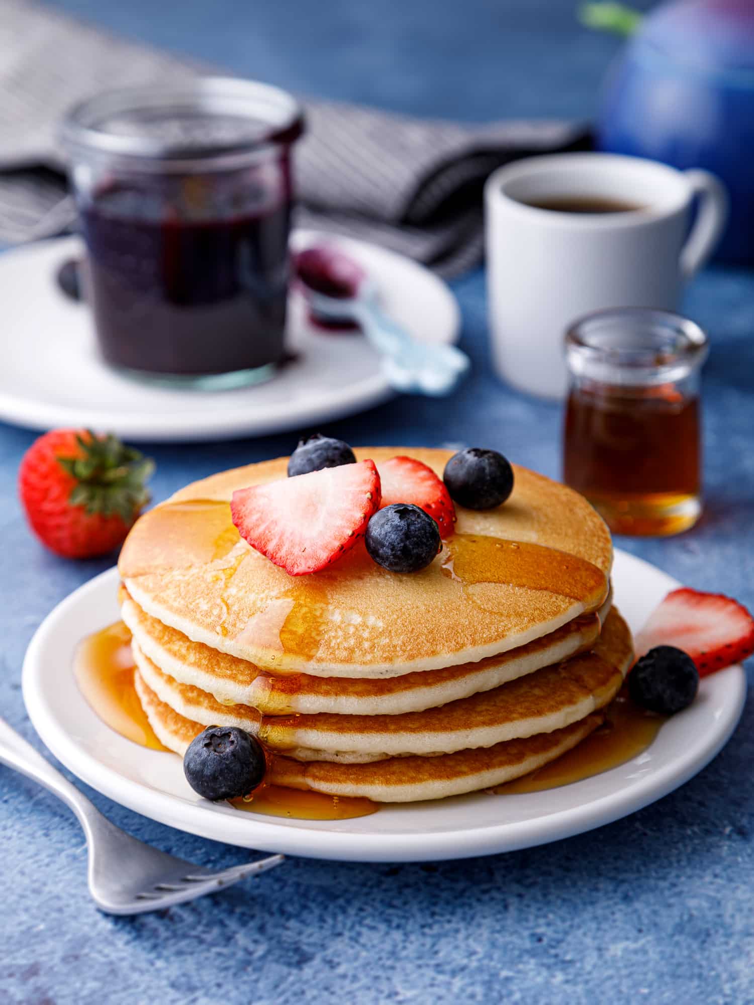 Stack of almond milk pancakes on a table with syrup, blueberry compote and a mug of hot tea on a table with a strawberry