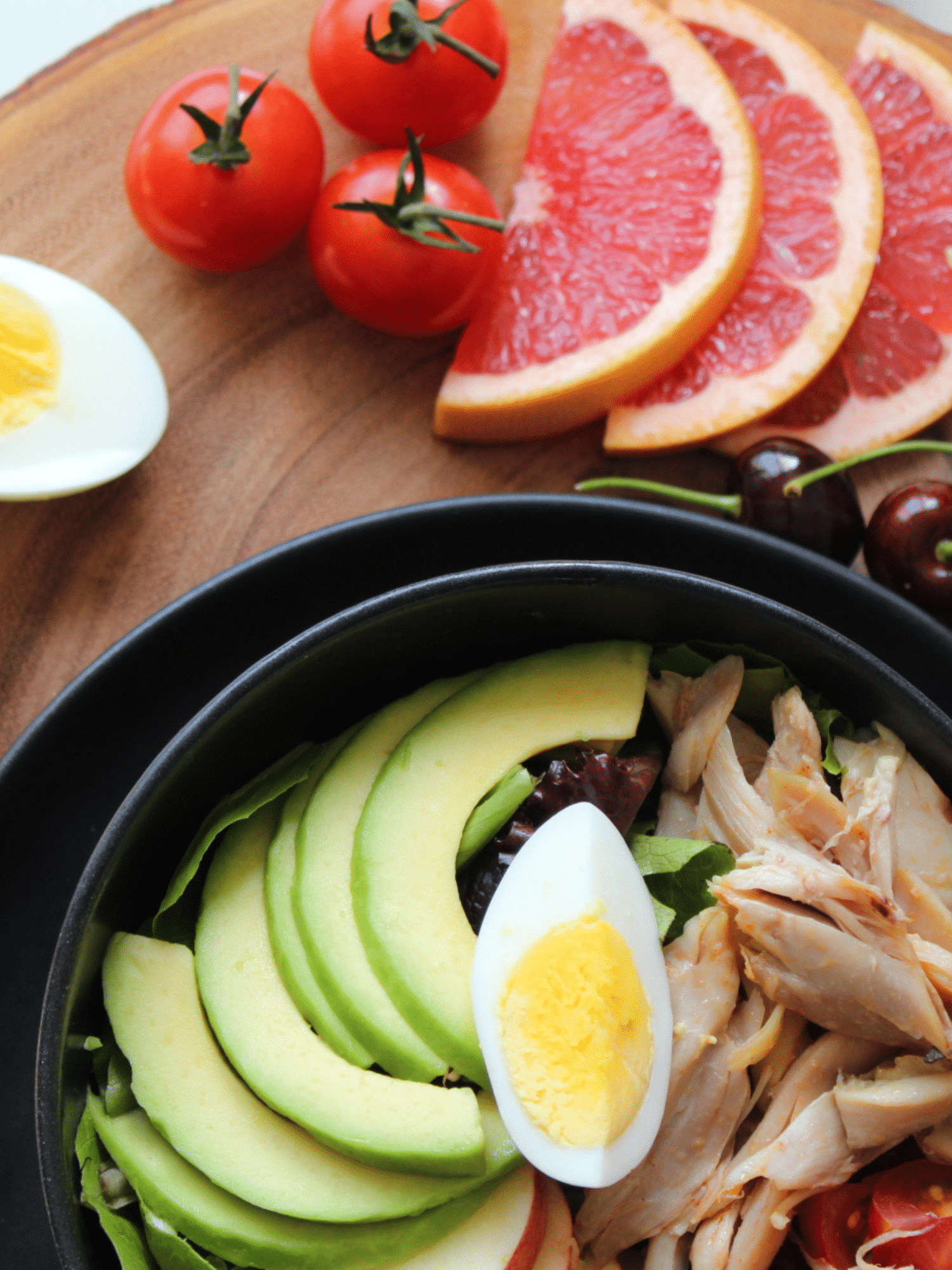 bowl of shredded chicken with slices of avacado and a slice of boiled egg on a table with sliced grapefruit, cherry tomatoes and half a boiled egg