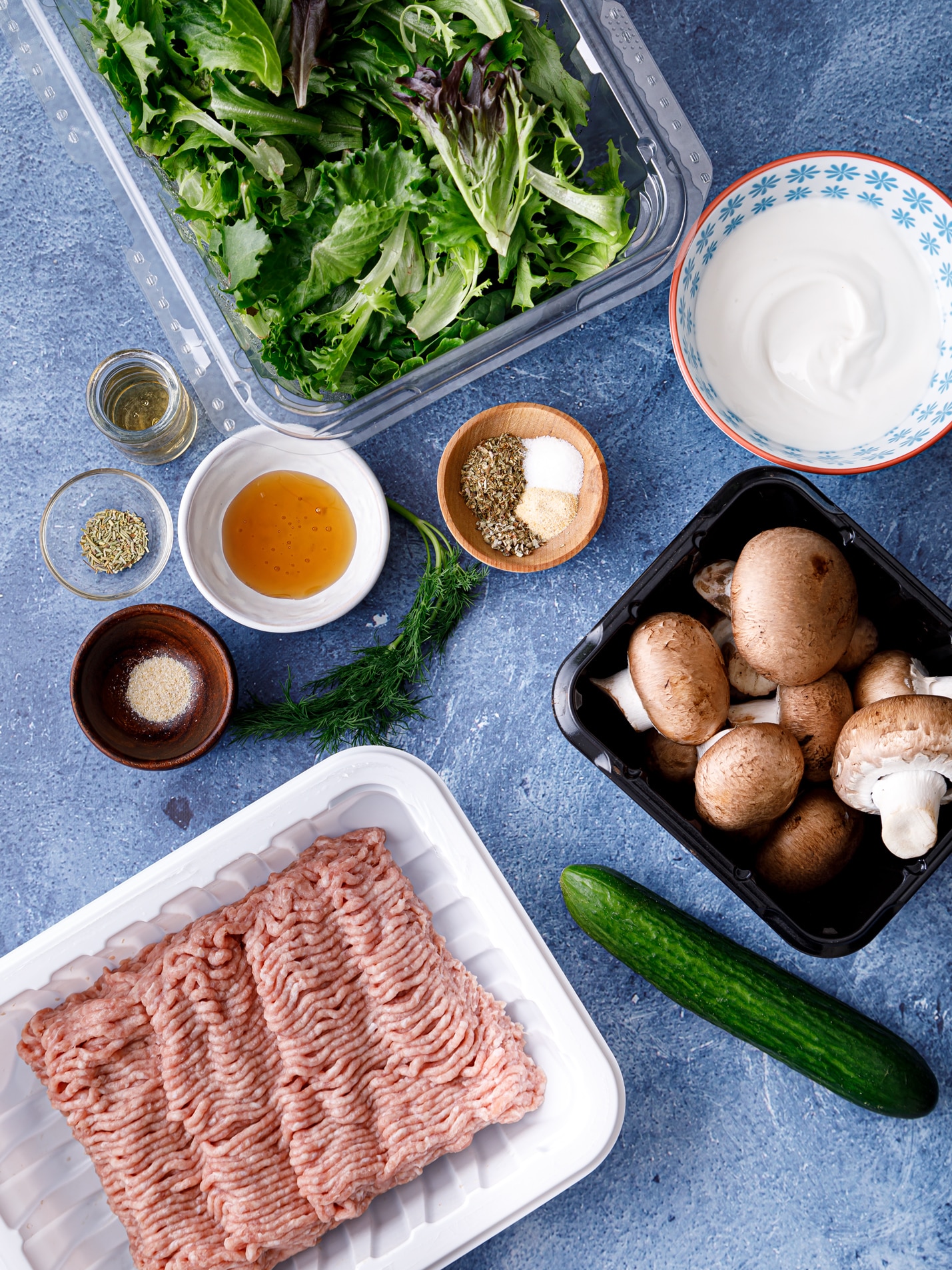 Groud turkey, a cucumber, fresh mushrooms, oregano, lettuce, a bowl of honey all sit on a blue table