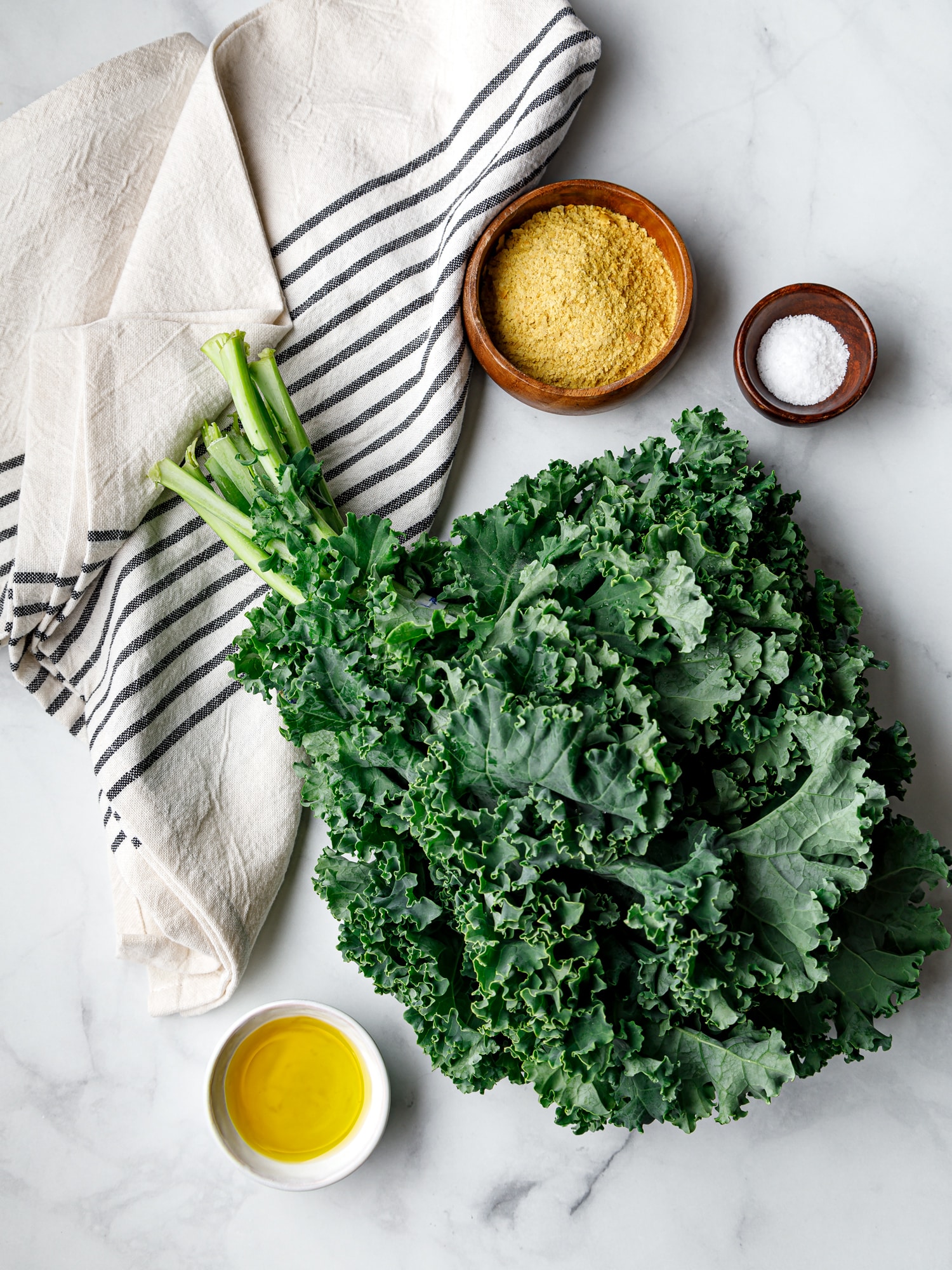 Fresh Kale with a bowl of salt and bowl of nutritional yeast and a bowl of avacado oil on a marble tabletop with a striped dishcloth