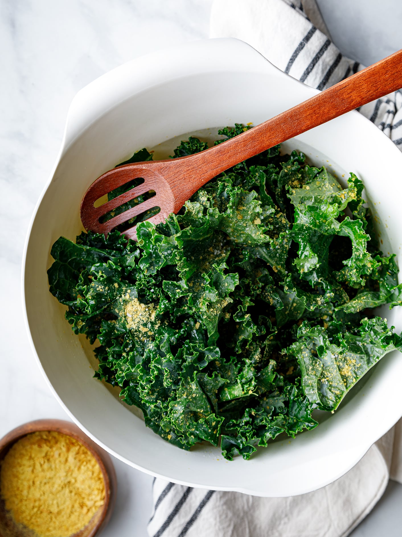 white bowl of raw kale sprinkled with seasonings with a wooden spoon for mixing and a small bowl of nutritional yeast on the side