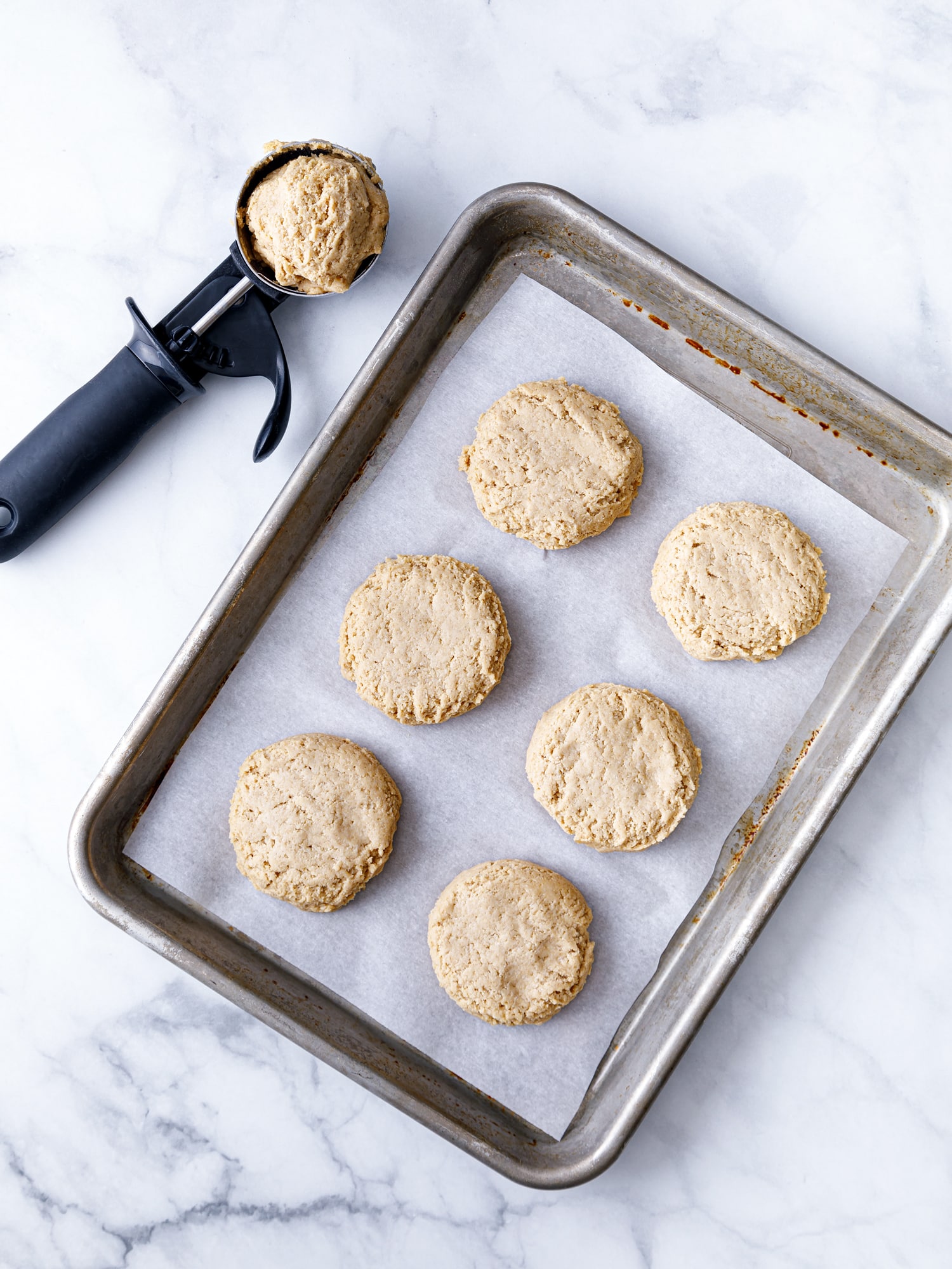 baking sheet covered in parchment paper with 6 drop biscuits padded out with a cookie scoop next to the tray