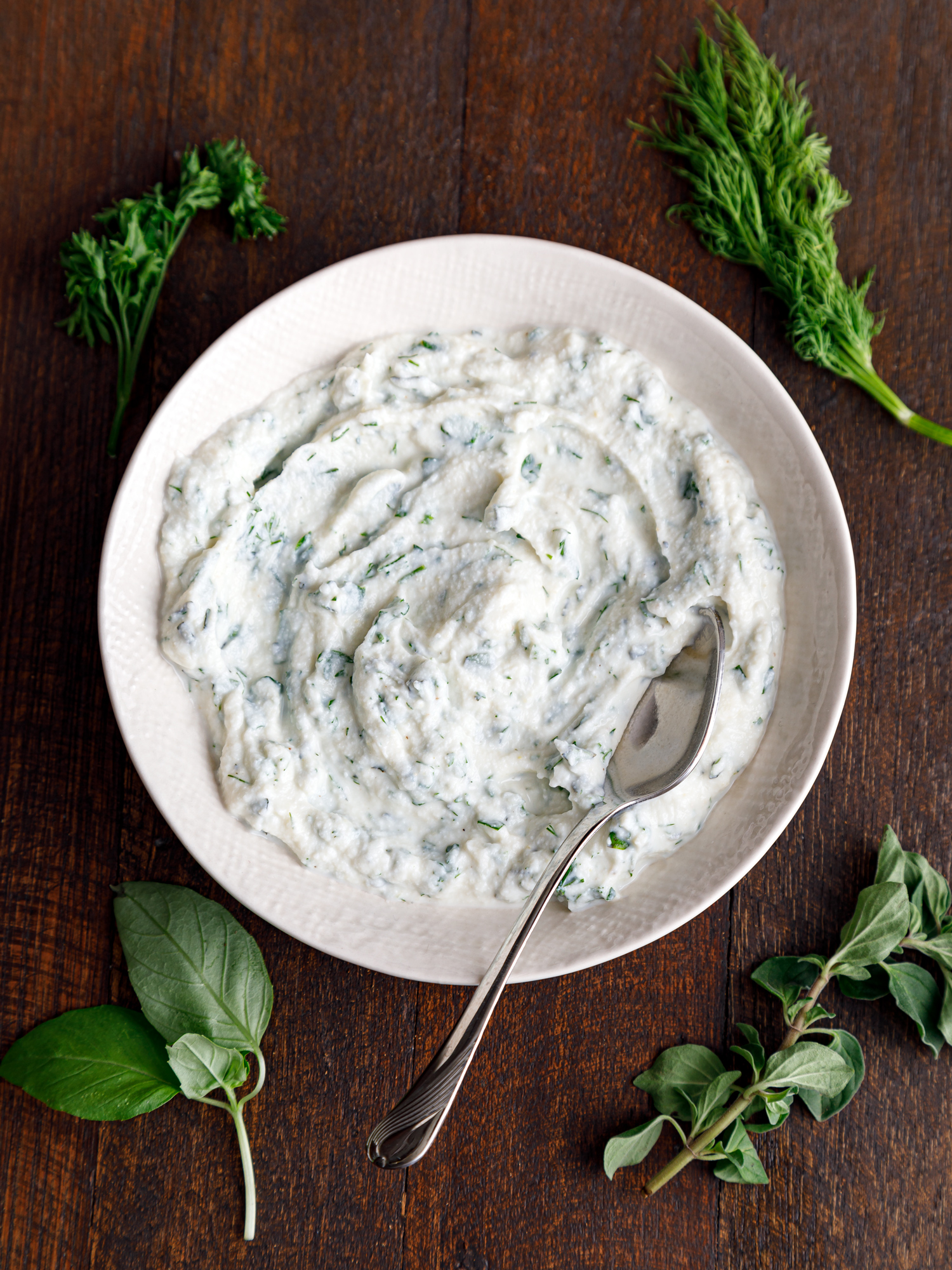 a white bowl of herbed ricotta cheese on a table surrounded by fresh herbs like basil, dill and parsley