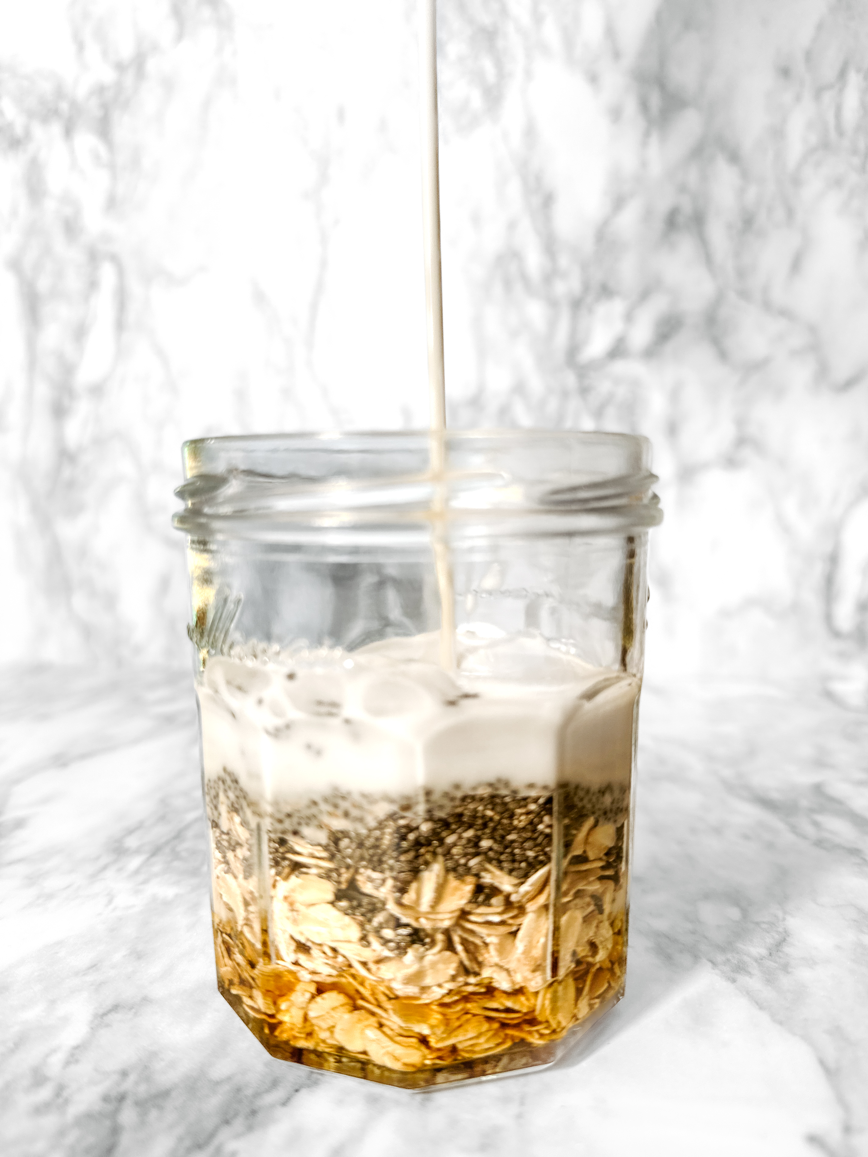 Jar of gluten free oats with chia seeds and maple syrup and coconut milk being poured over to make coconut milk overnight oats