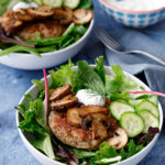 two deconstructed burger bowls consisting of two white bowls filled with fresh greens a turkey burger, sauteed mushrooms and a homemade quick pickle with a bowl of a tzatziki sauce