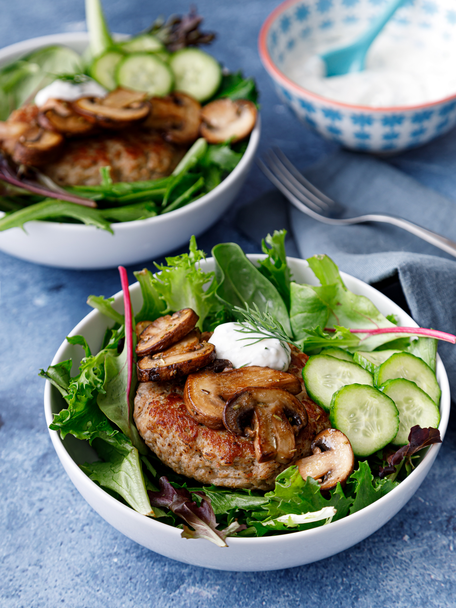 two deconstructed burger bowls consisting of two white bowls filled with fresh greens a turkey burger, sauteed mushrooms and a homemade quick pickle with a bowl of a tzatziki sauce