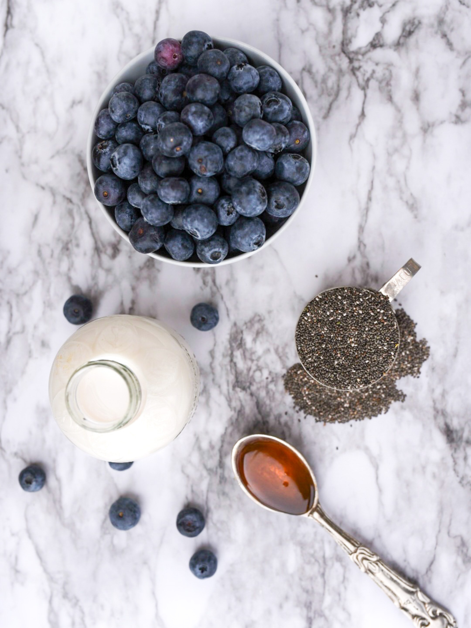 a marble table top with a bowl of blueberries, a measuring cup of chia seeds, a small jar of almond milk and a tablespoon of honey 