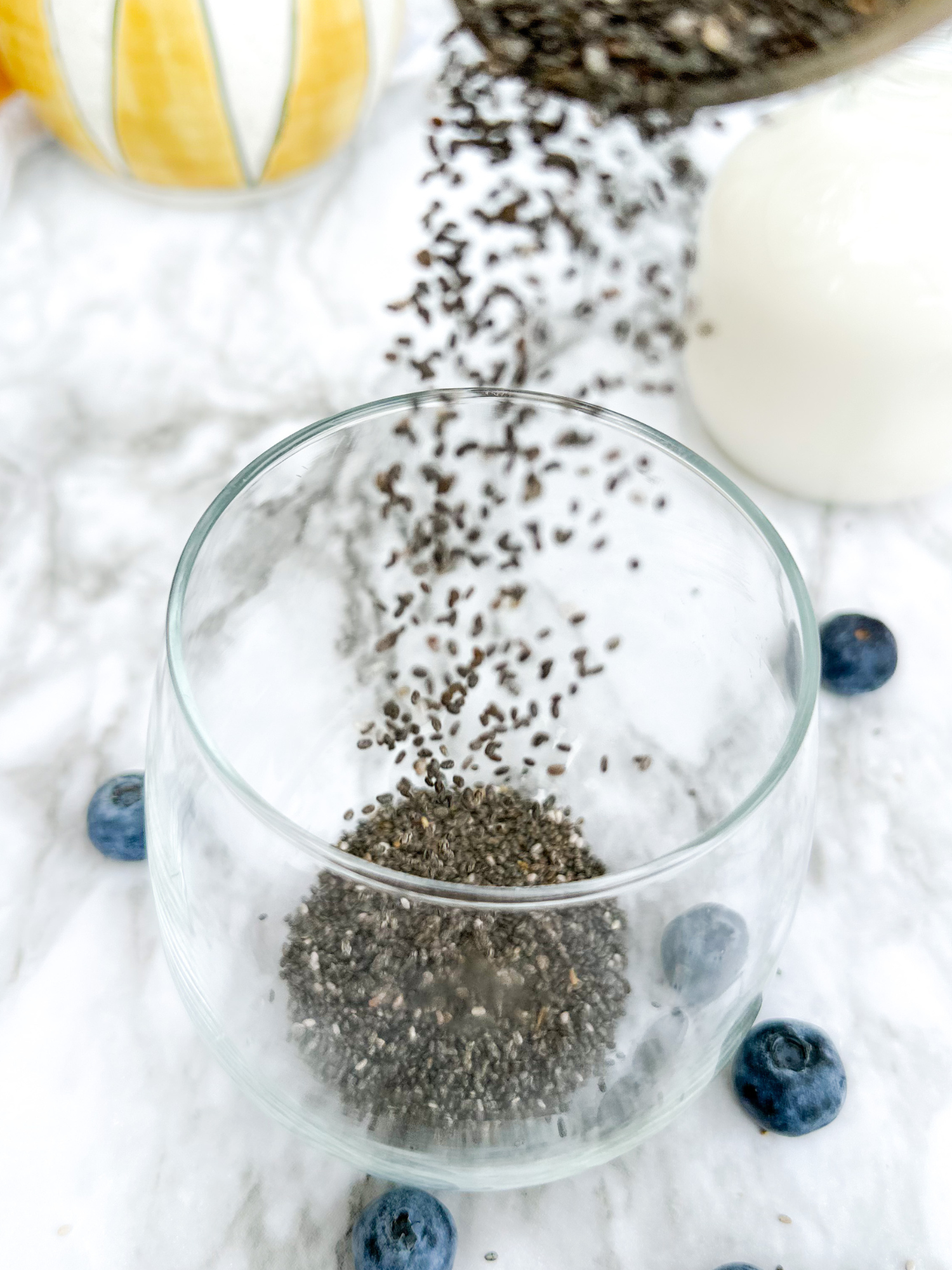 a glass cup on a table surrounded by blueberries with chia seeds being poured in