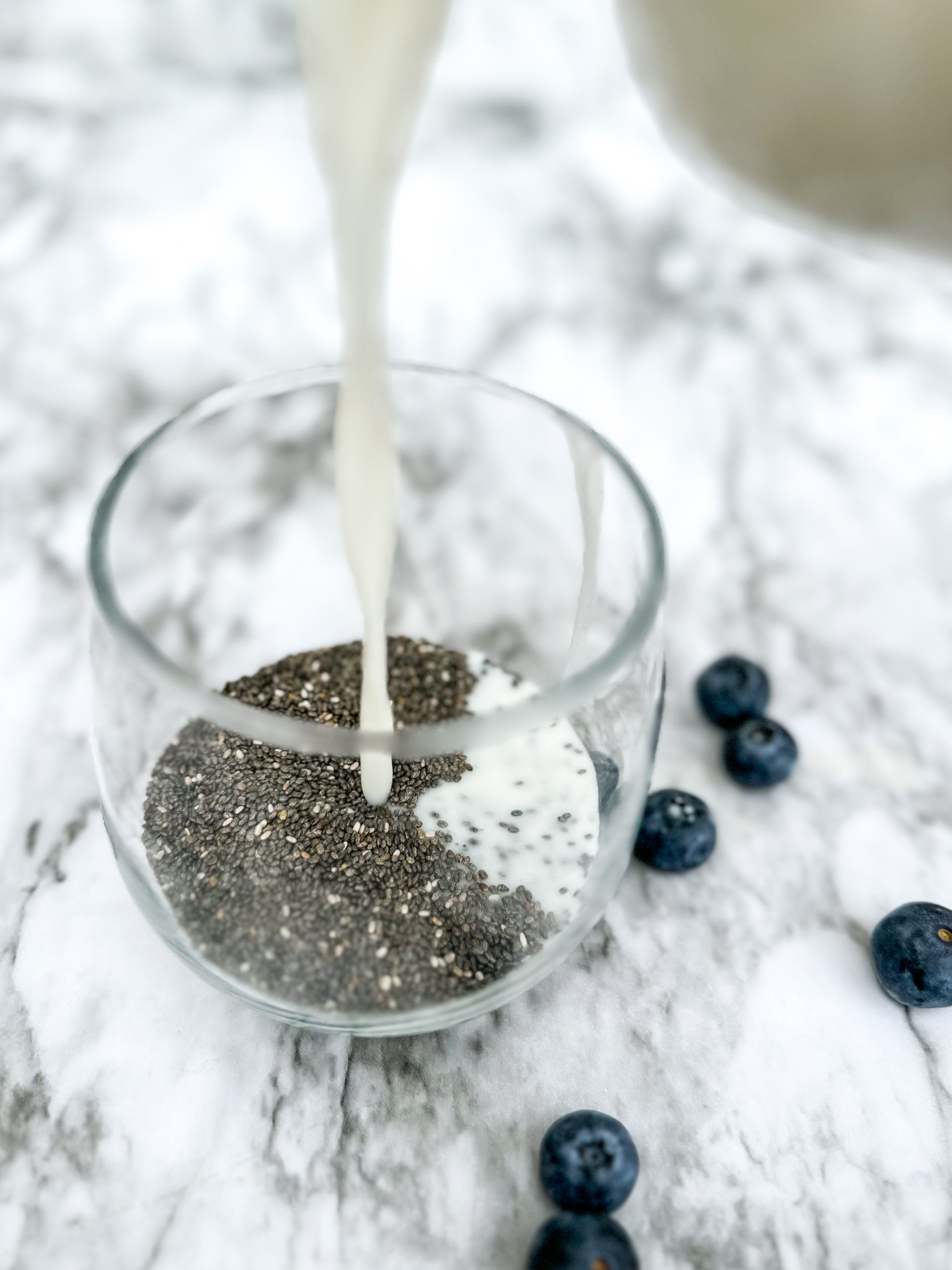 a marble tabletop with a glass cup with chia seeds at the bottom and almond milk being poured in