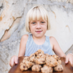 a little blond girl holding out a wooden tray stacked with gluten free dairy free cookies