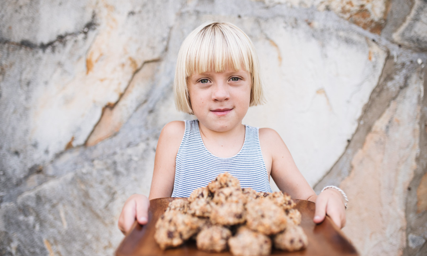 a little blond girl holding out a wooden tray stacked with gluten free dairy free cookies