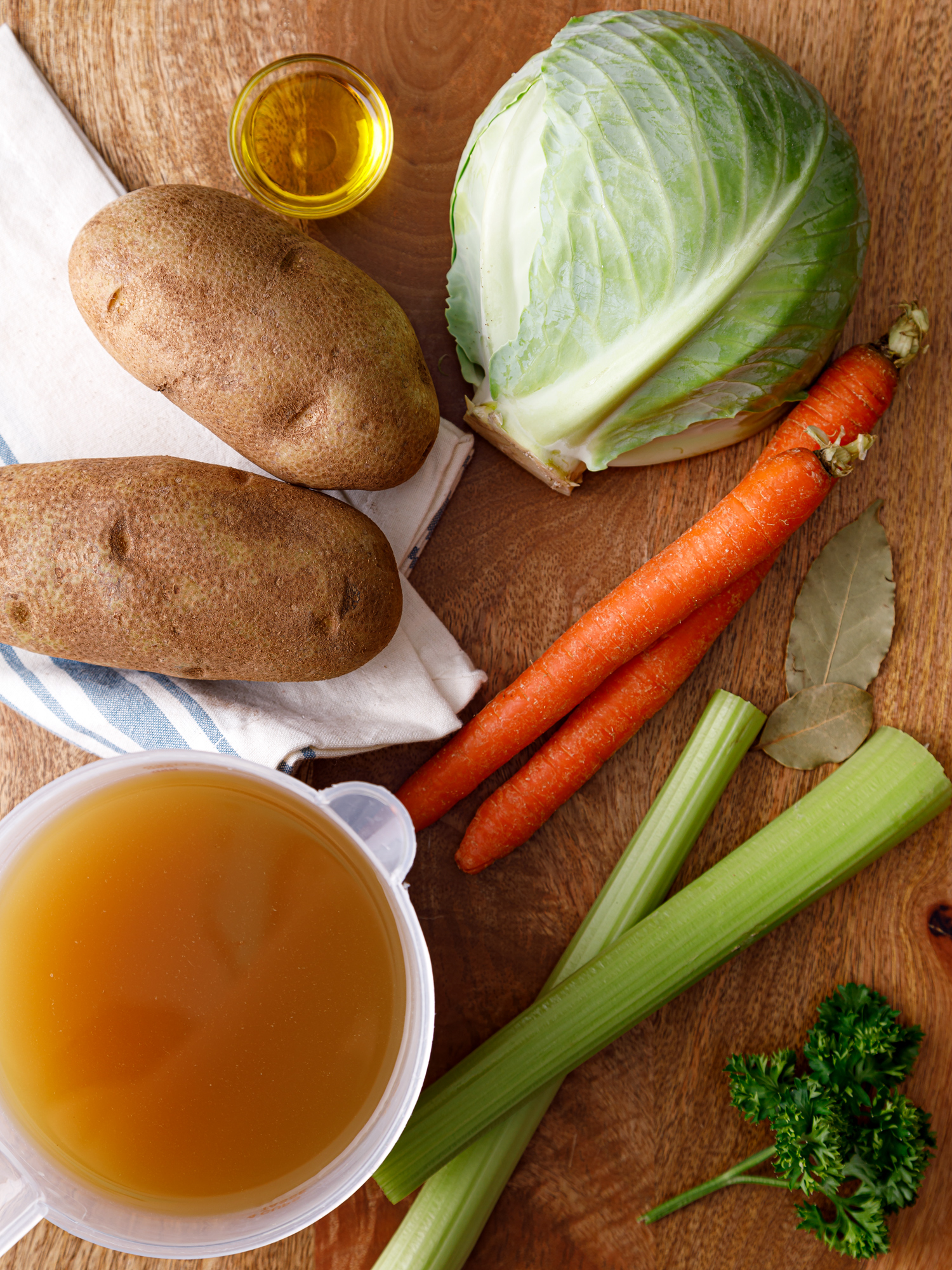 two potaotes, a green cabbage, two carrots, two celery sticks, some parsely and measuring cup of bone broth laid out on a table