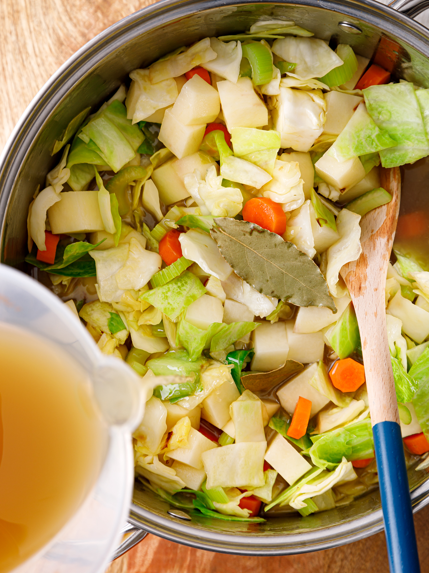 a pot of chopped cabbage, carrots, celery and potatoes with bone broth being poured in and stirred with a wooden spoon