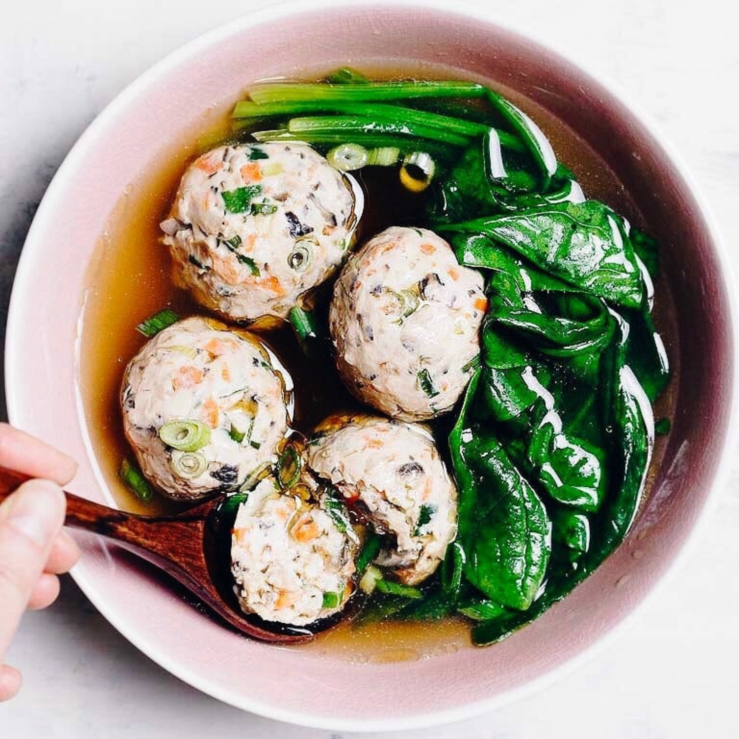 a pink bowl of asian chicken meatball soup with spinach and a hand holding a spoon