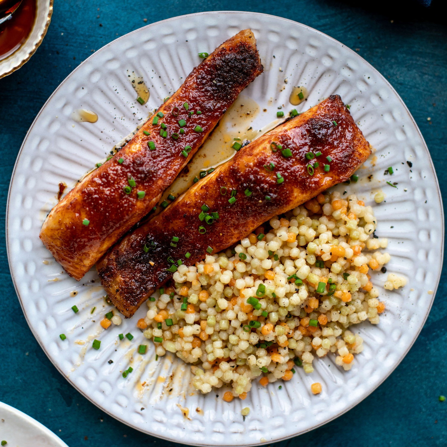 a white plate on a blue table with two pieces of maple bbq salmon next to a serving of brown butter couscous