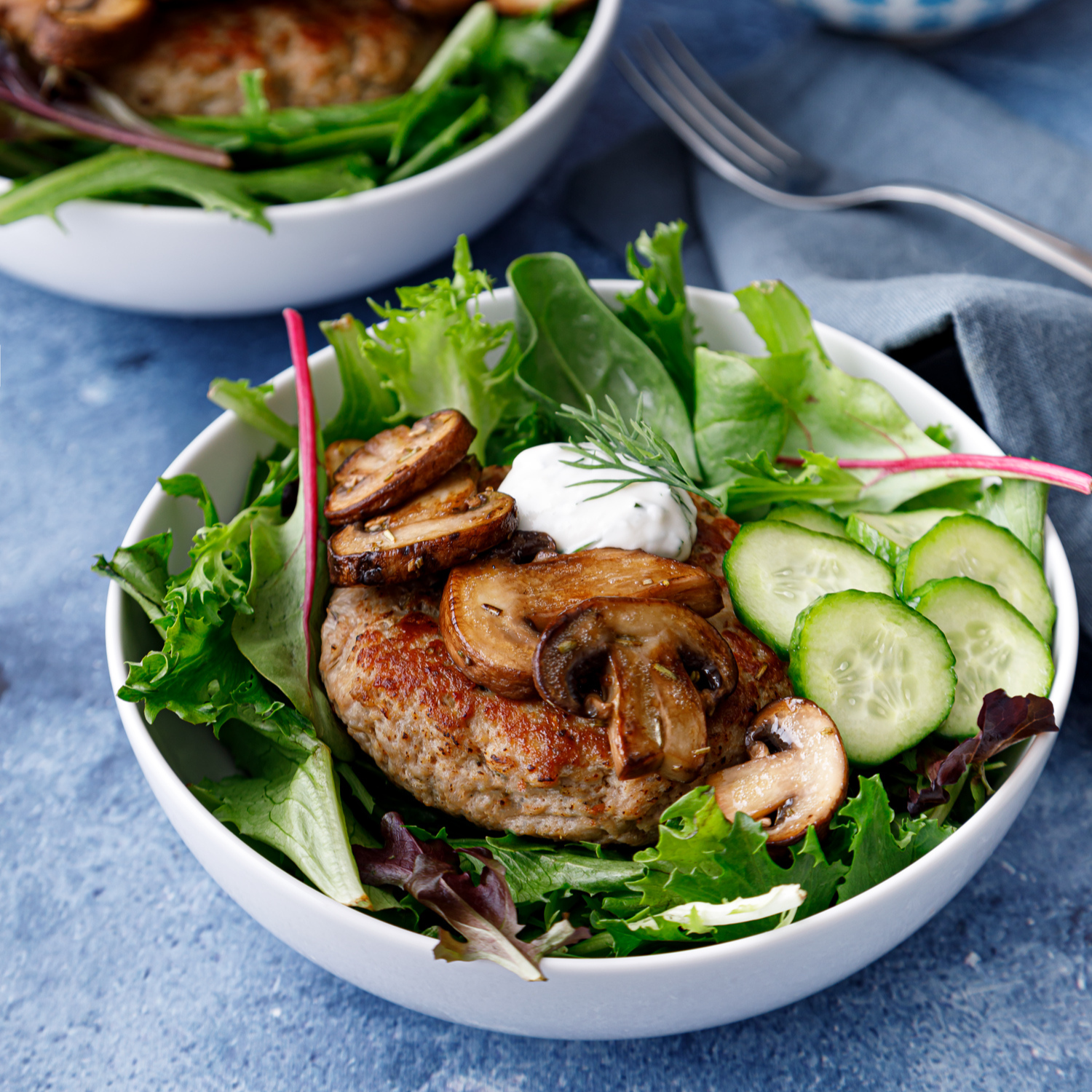 two deconstructed burger bowls consisting of two white bowls filled with fresh greens a turkey burger, sauteed mushrooms and a homemade quick pickle with a bowl of a tzatziki sauce