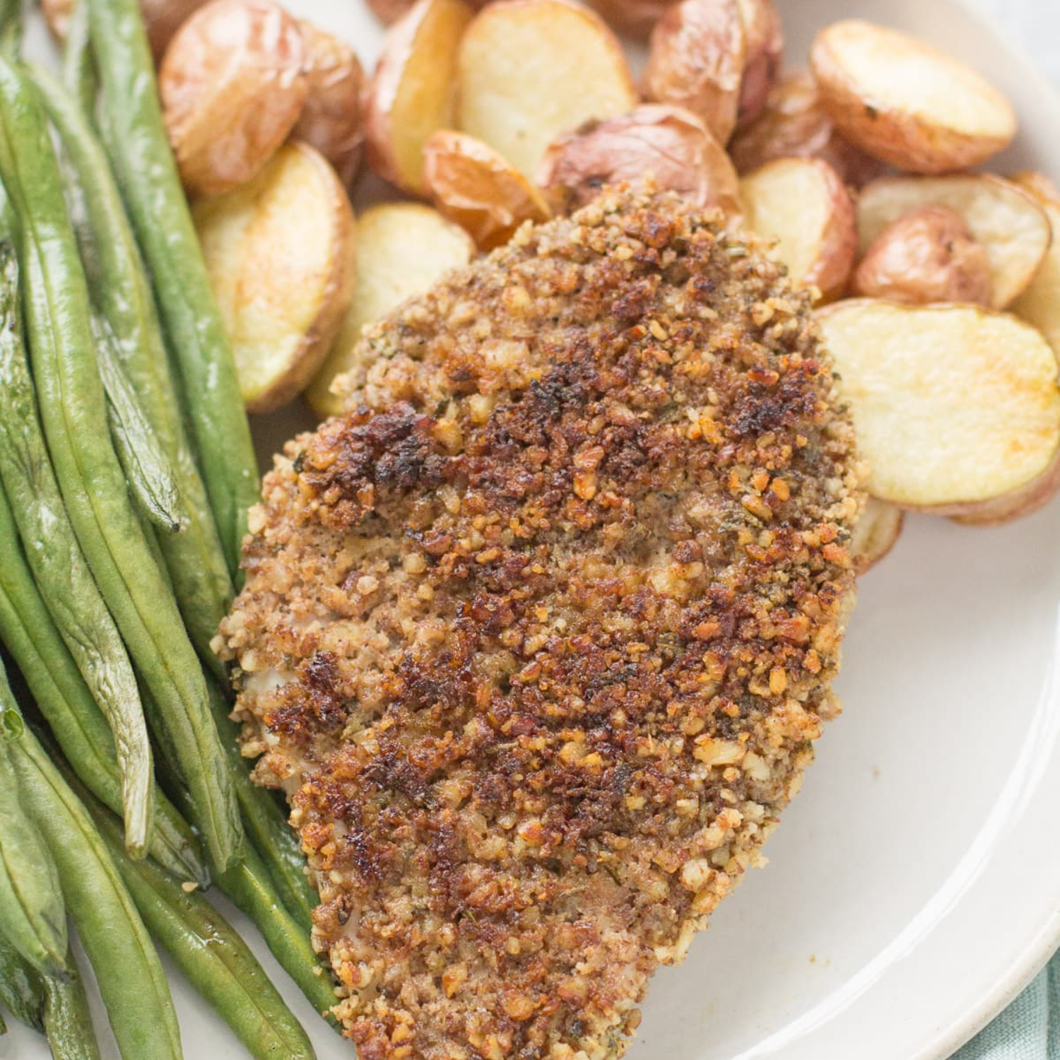 A piece of pecan roasted pork chops next to green beans and some roasted potatoes on a white plate