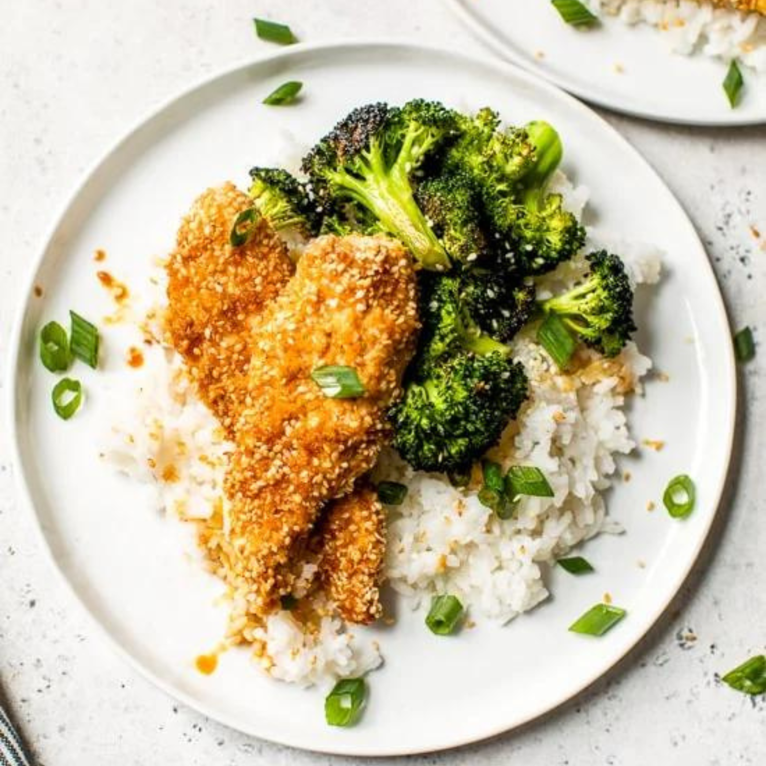 a white plate with two pieces of sesame encrusted chicken tenders with a side of roasted broccoli and jasmine rice