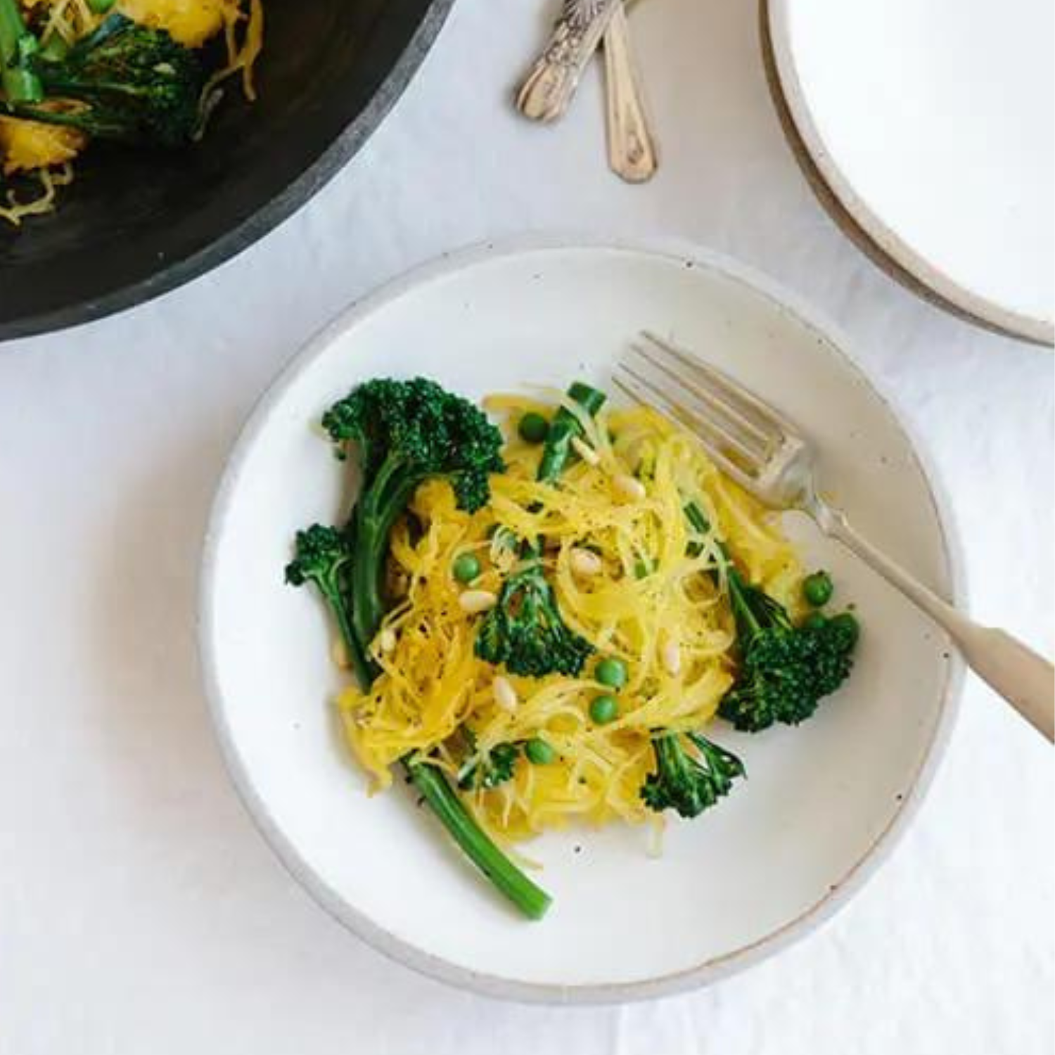 a white bowl of roasted spaghetti squash with pieces of broccolini on a white table with silverware