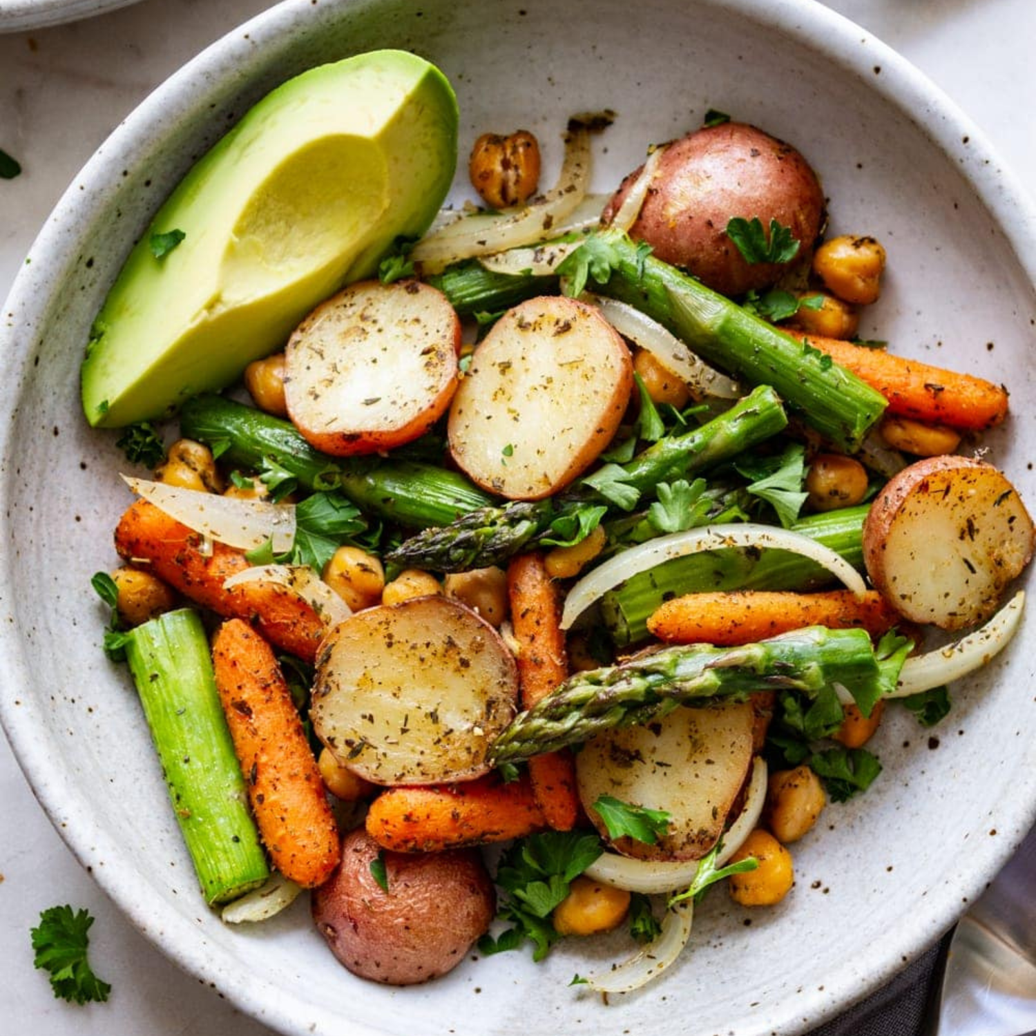 A white bowl with roasted asparagus, carrots, potatoes and chickpeas with a slice of avacado 