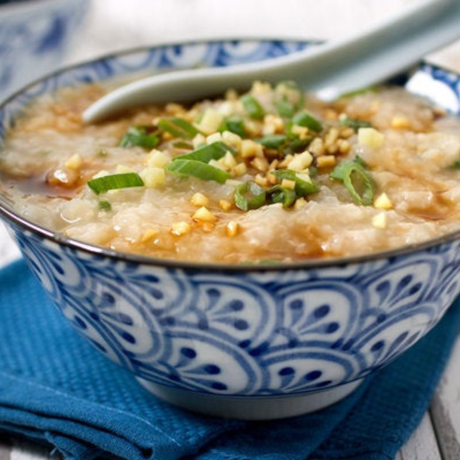 a blue and white bowl on a table filled with chicken congee and topped with chopped scallions with a large white spoon