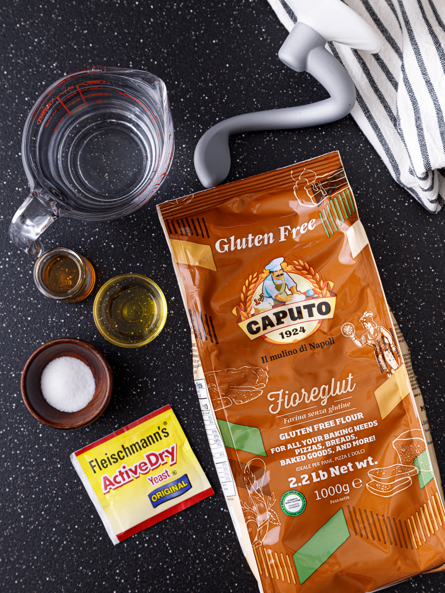 Gluten Free flour, yeast, salt, oil and a cup of warm water with a dough hook on a dark table top